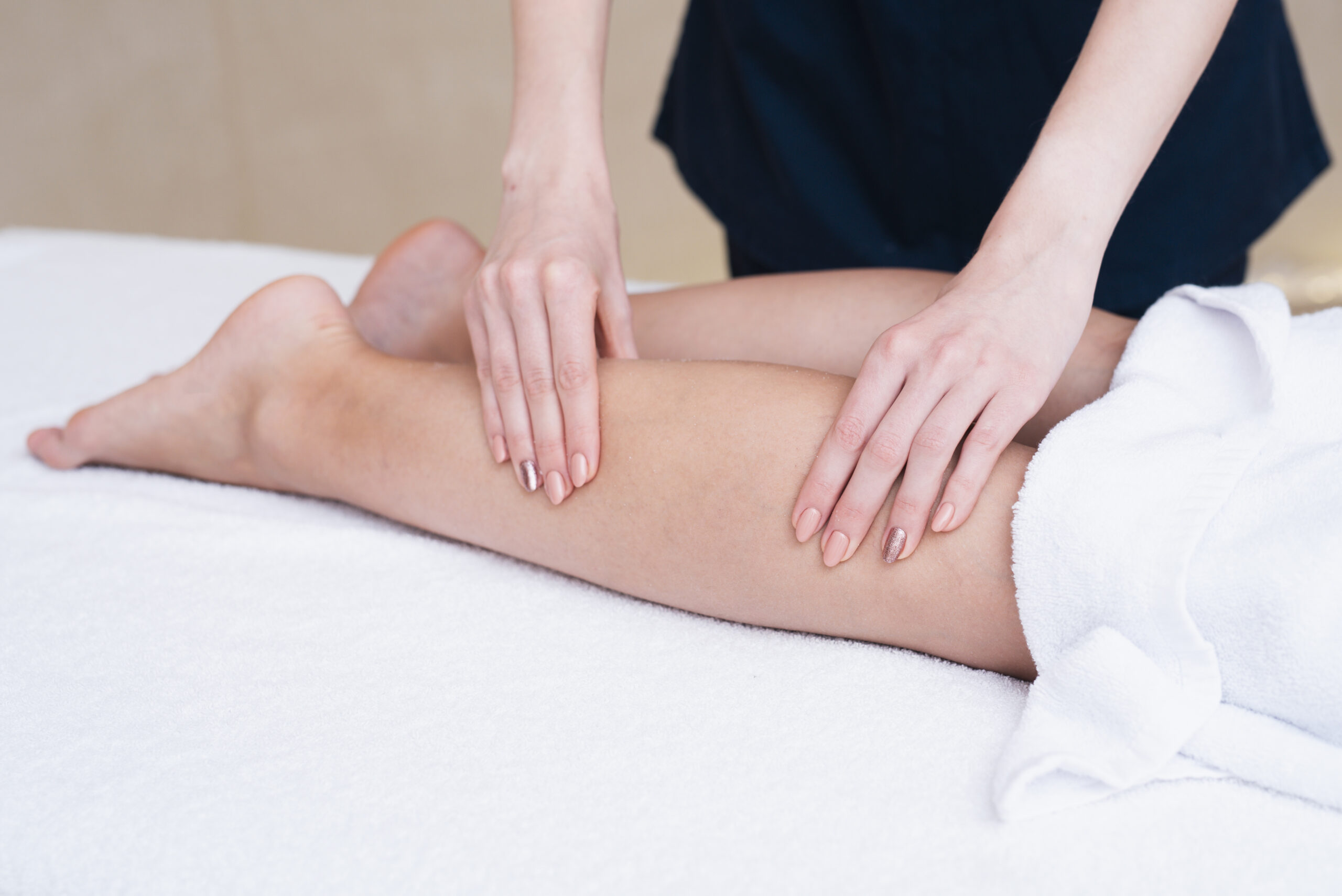 Terapeuta wykonuje masaż nóg pacjentki na białym stole, w tle jasne, neutralne tło.