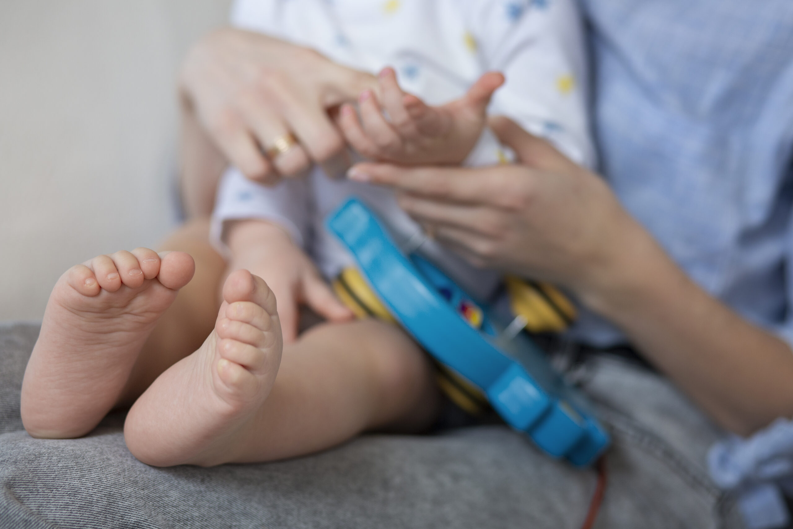 Dziecko leży na kolanach dorosłej osoby, trzyma ją za rękę, a na stopach ma aparat ortopedyczny.