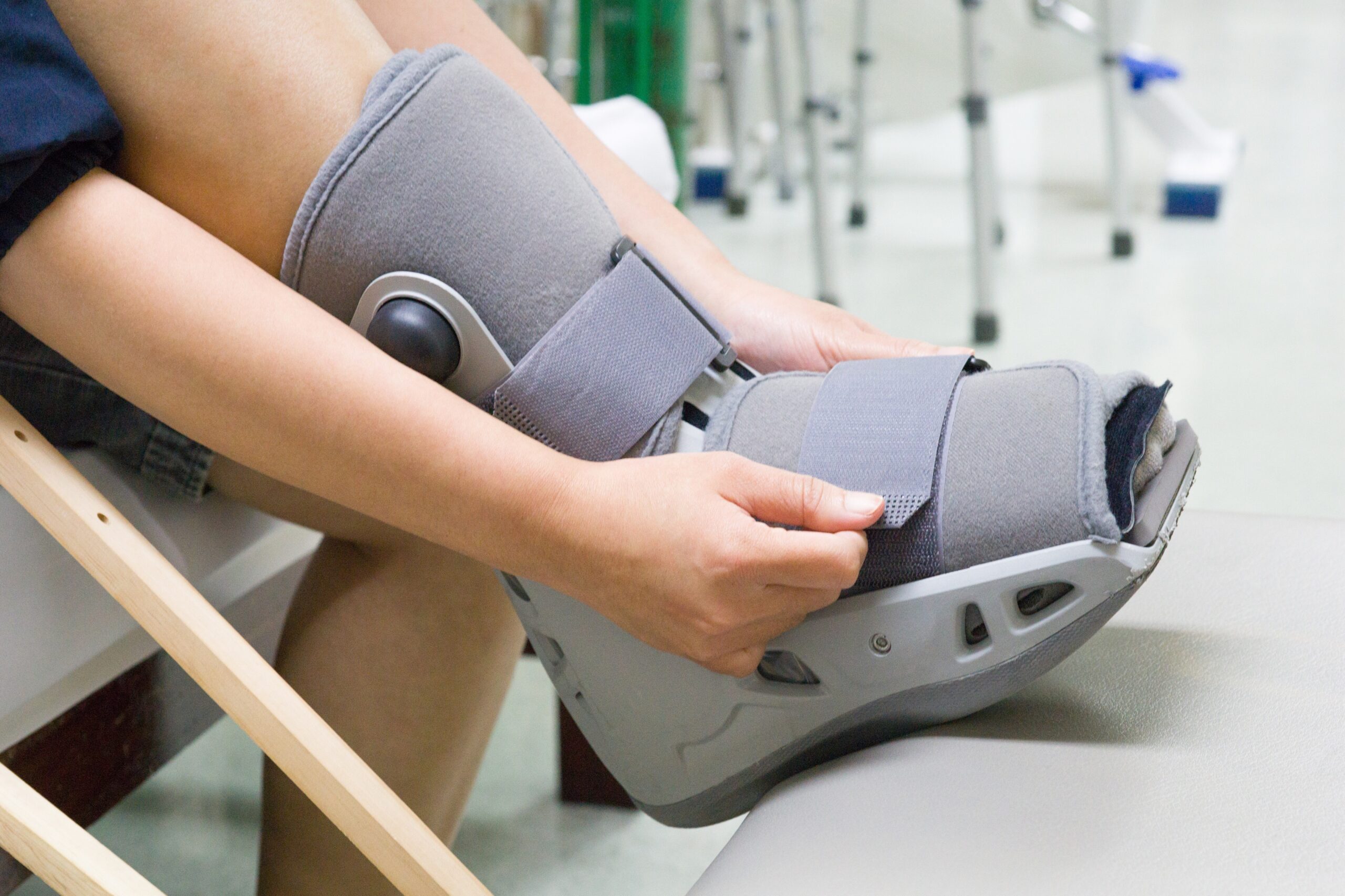 Osoba zakłada stabilizator na nogę w warunkach rehabilitacyjnych, w tle sprzęt medyczny i łóżko.