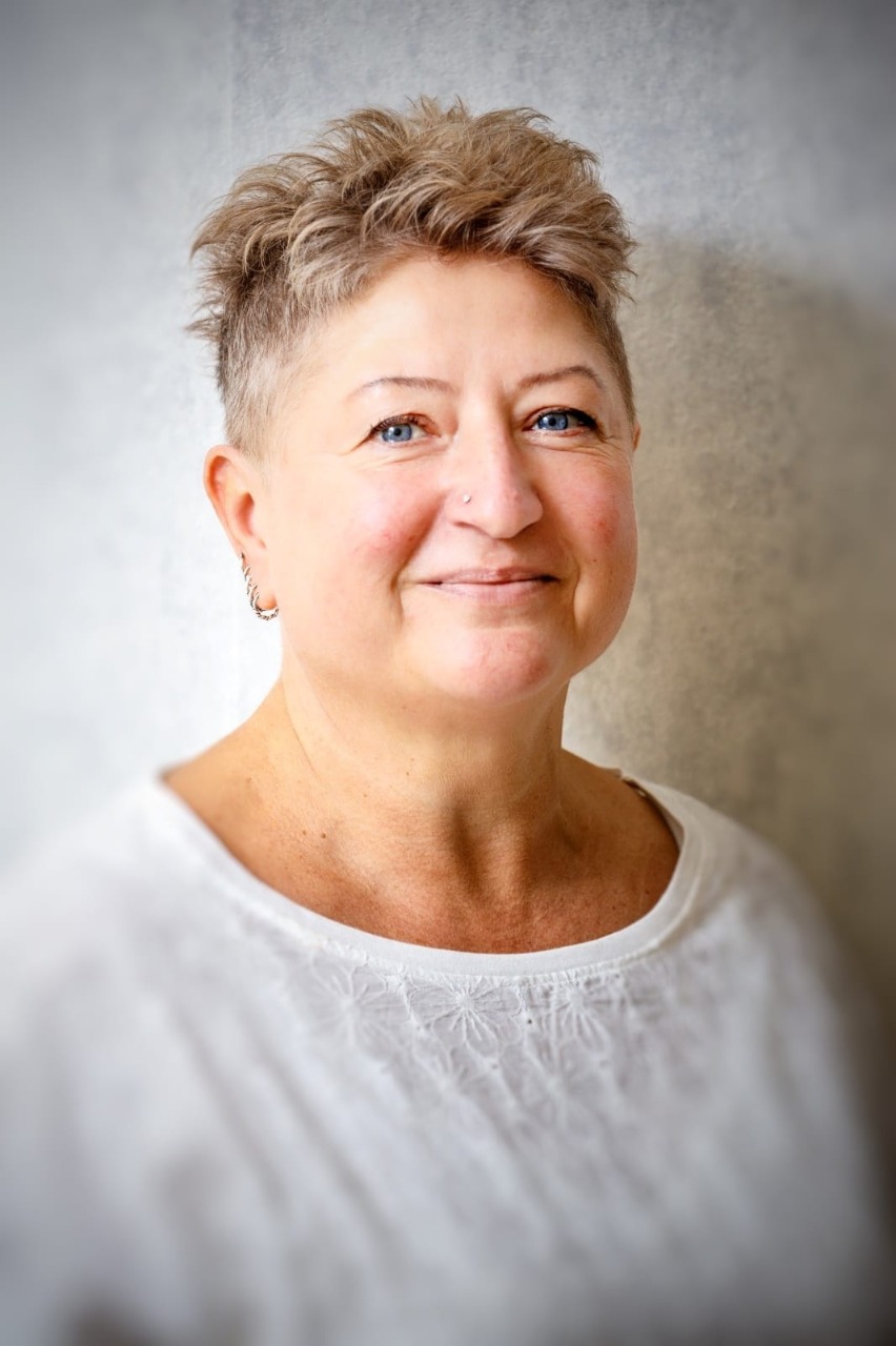 Uśmiechnięta kobieta z krótkimi, kręconymi włosami, nosząca białą bluzkę, na tle jasnej, gładkiej ściany.