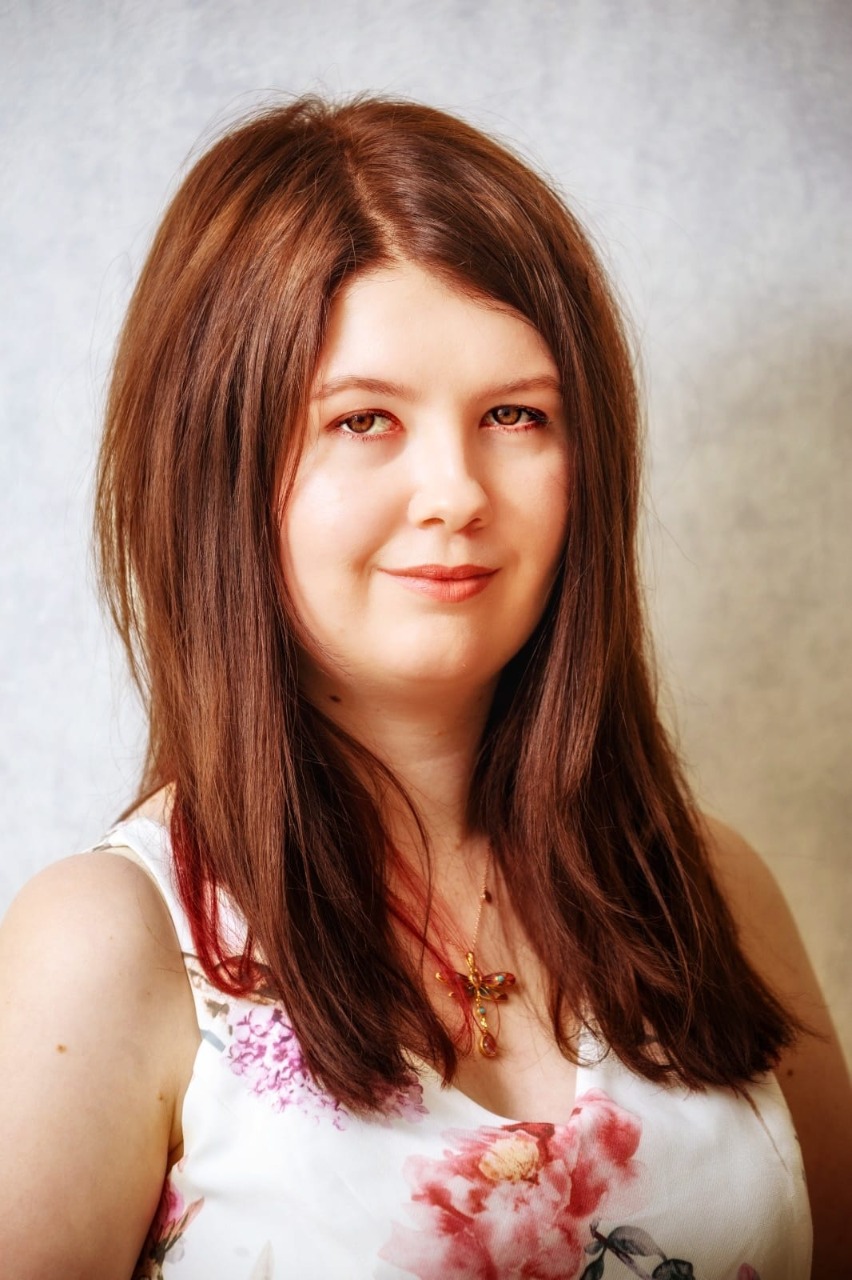 Kobieta z długimi brązowymi włosami, uśmiechnięta, w białej bluzce z różowymi kwiatami, na tle jasnej ściany.