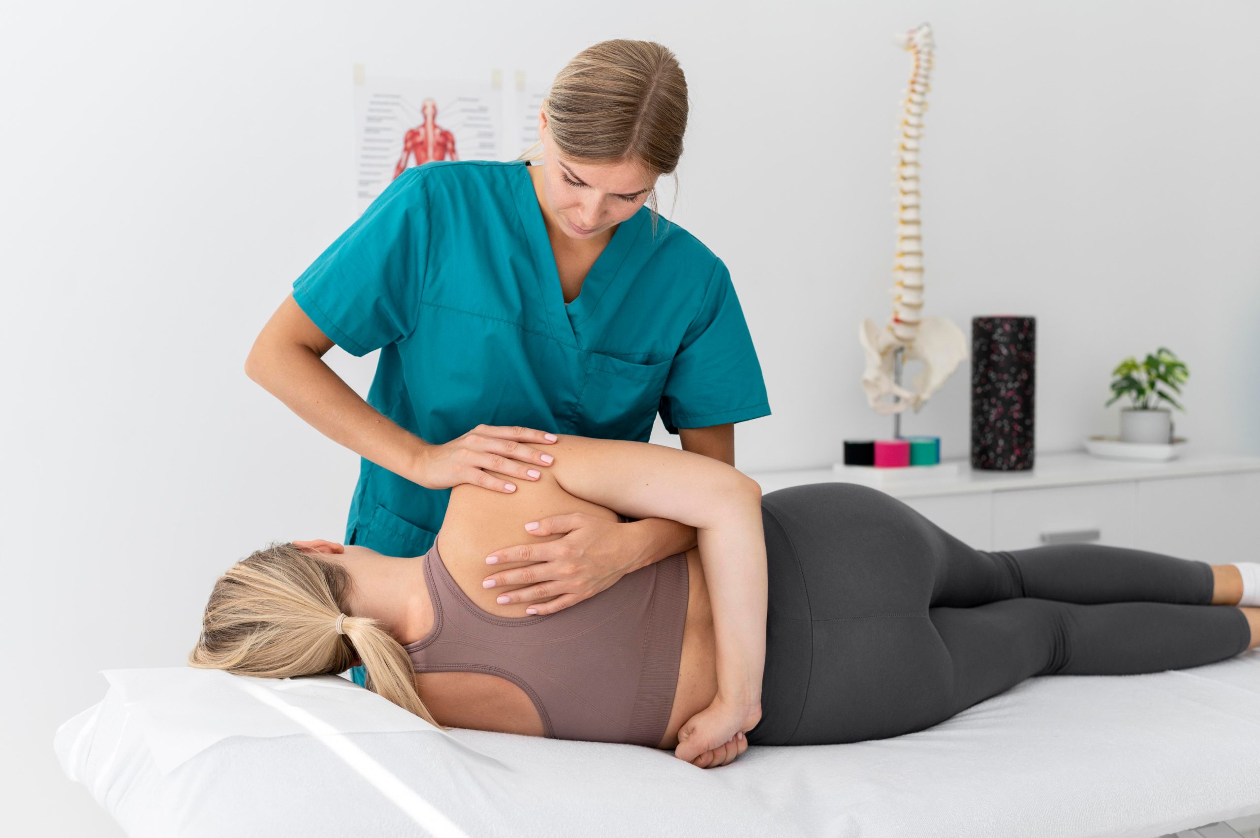 Fizjoterapeuta wykonuje masaż pleców kobiety leżącej na stole w gabinecie z modelami kręgosłupa i mięśni na ścianie.