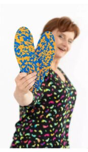 Kobieta trzyma dwie buty w stylu espadryli z niebiesko-żółtym wzorem, uśmiecha się i patrzy w stronę kamery.