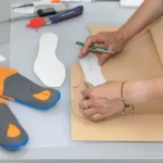 Osoba wycina wzór na papierze, obok leżą buty sportowe z miękką podeszwą i narzędzia do szycia.