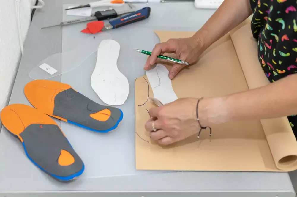 Osoba wycina wzór na papierze, obok leżą buty sportowe z miękką podeszwą i narzędzia do szycia.