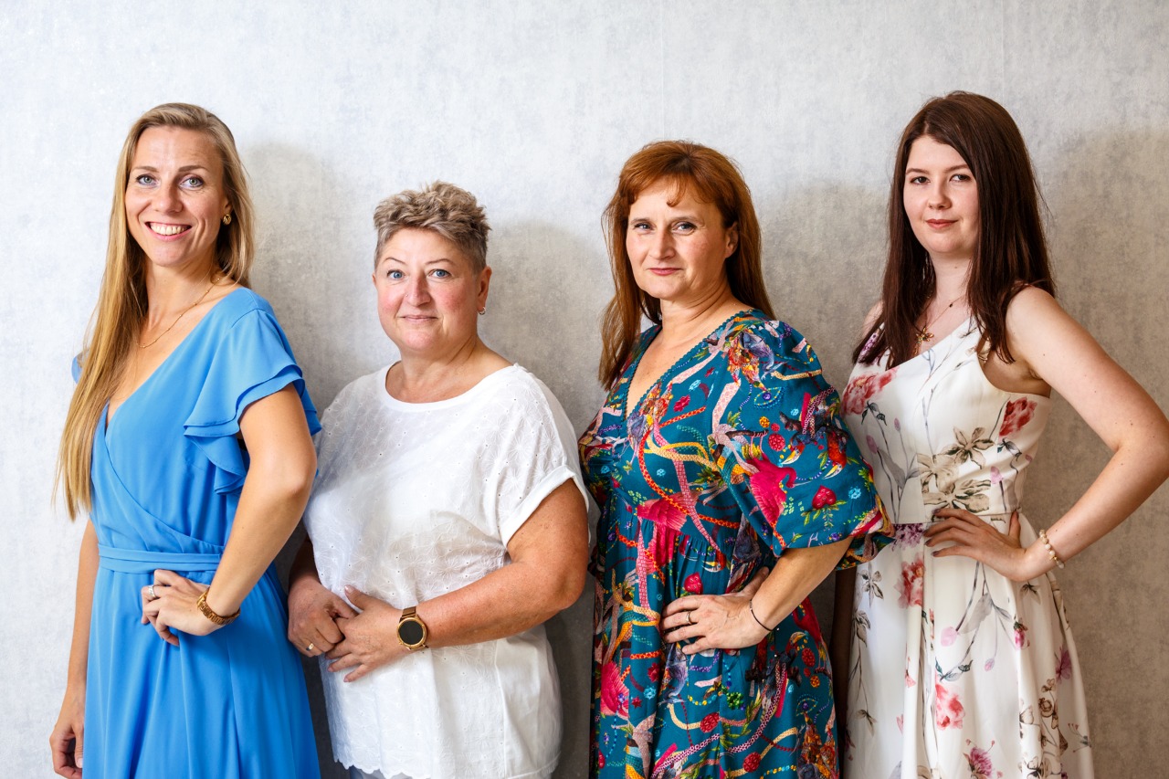 Cztery kobiety stojące obok siebie na tle jasnej ściany, ubrane w eleganckie, kolorowe sukienki, uśmiechnięte i patrzące