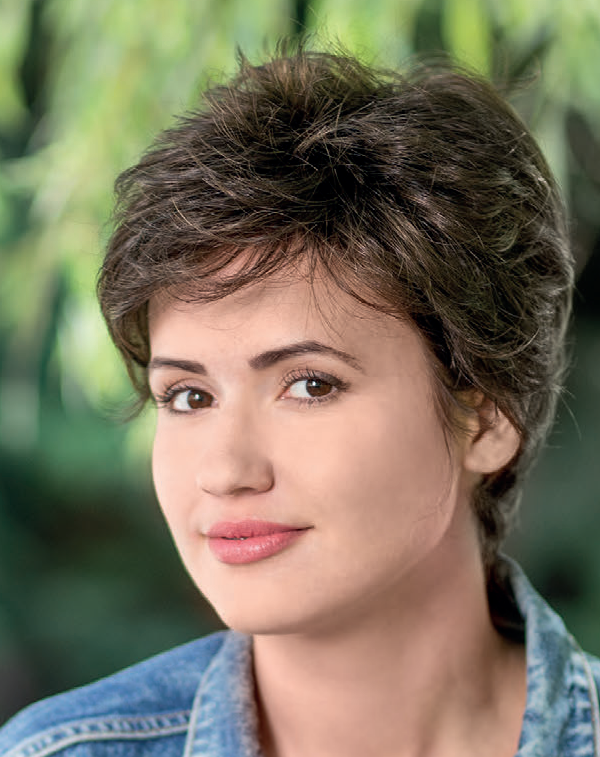 Młoda kobieta z krótkimi brązowymi włosami, uśmiechnięta, na tle zielonych liści.