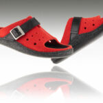 Czerwone kapcie z czarnymi paskami i metalowymi klamrami, uniesione nad lustrem, z widoczną refleksją na powierzchni.