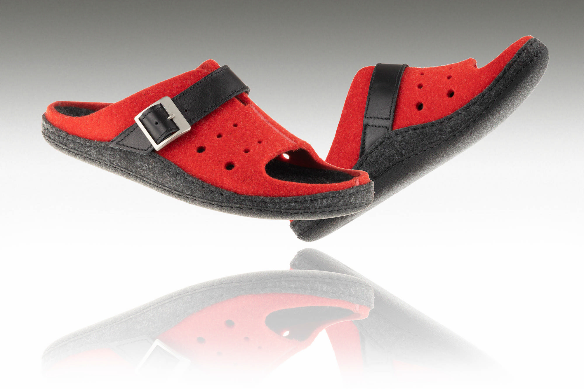 Czerwone kapcie z czarnymi paskami i metalowymi klamrami, uniesione nad lustrem, z widoczną refleksją na powierzchni.