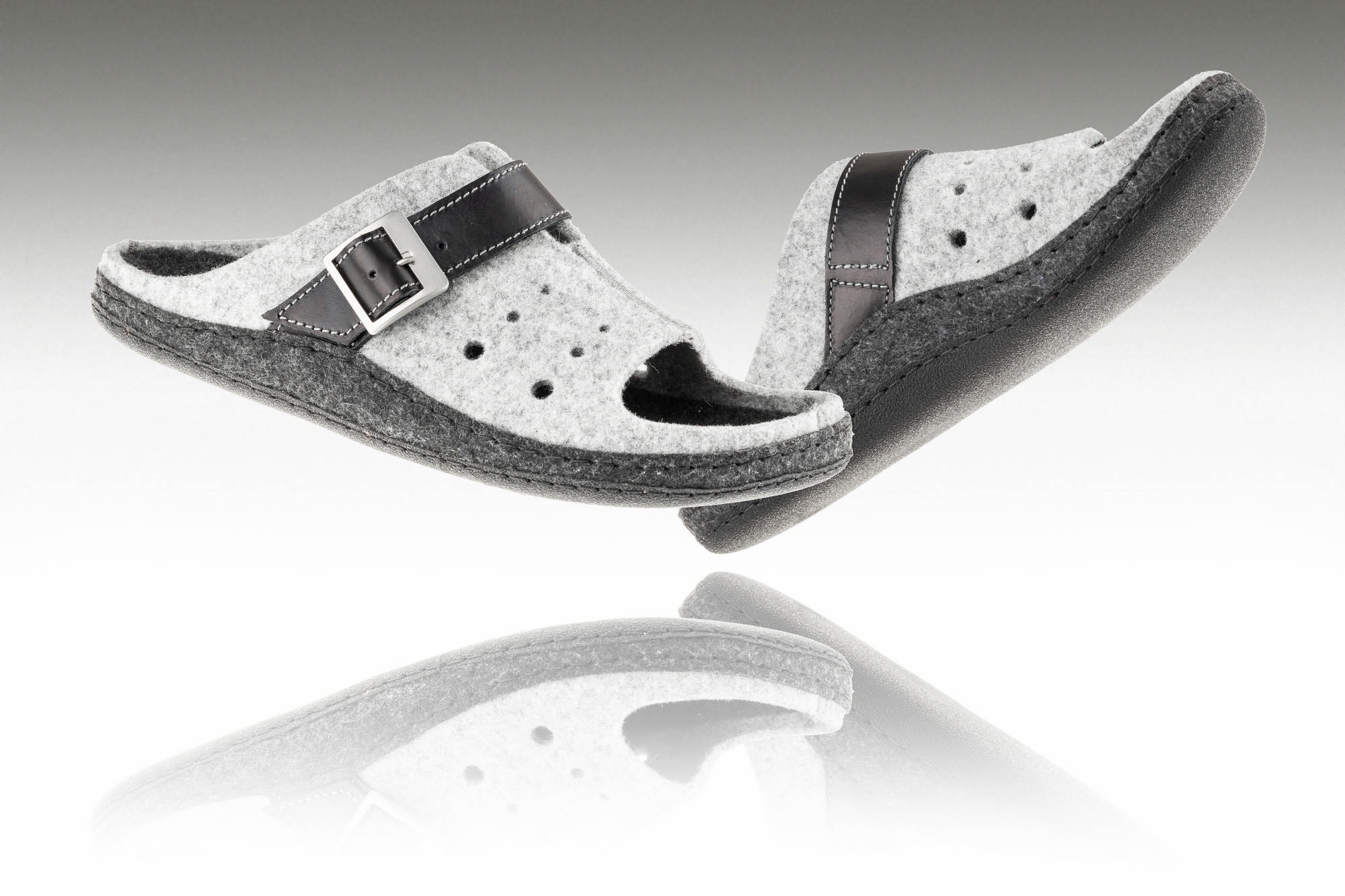 Kapcie z filcu i skóry z czarnymi paskami i metalową klamrą, uniesione nad odbiciem na białym tle.