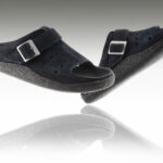 Czarne kapcie z dwoma paskami i metalowymi klamrami, uniesione nad odbiciem na białym tle.