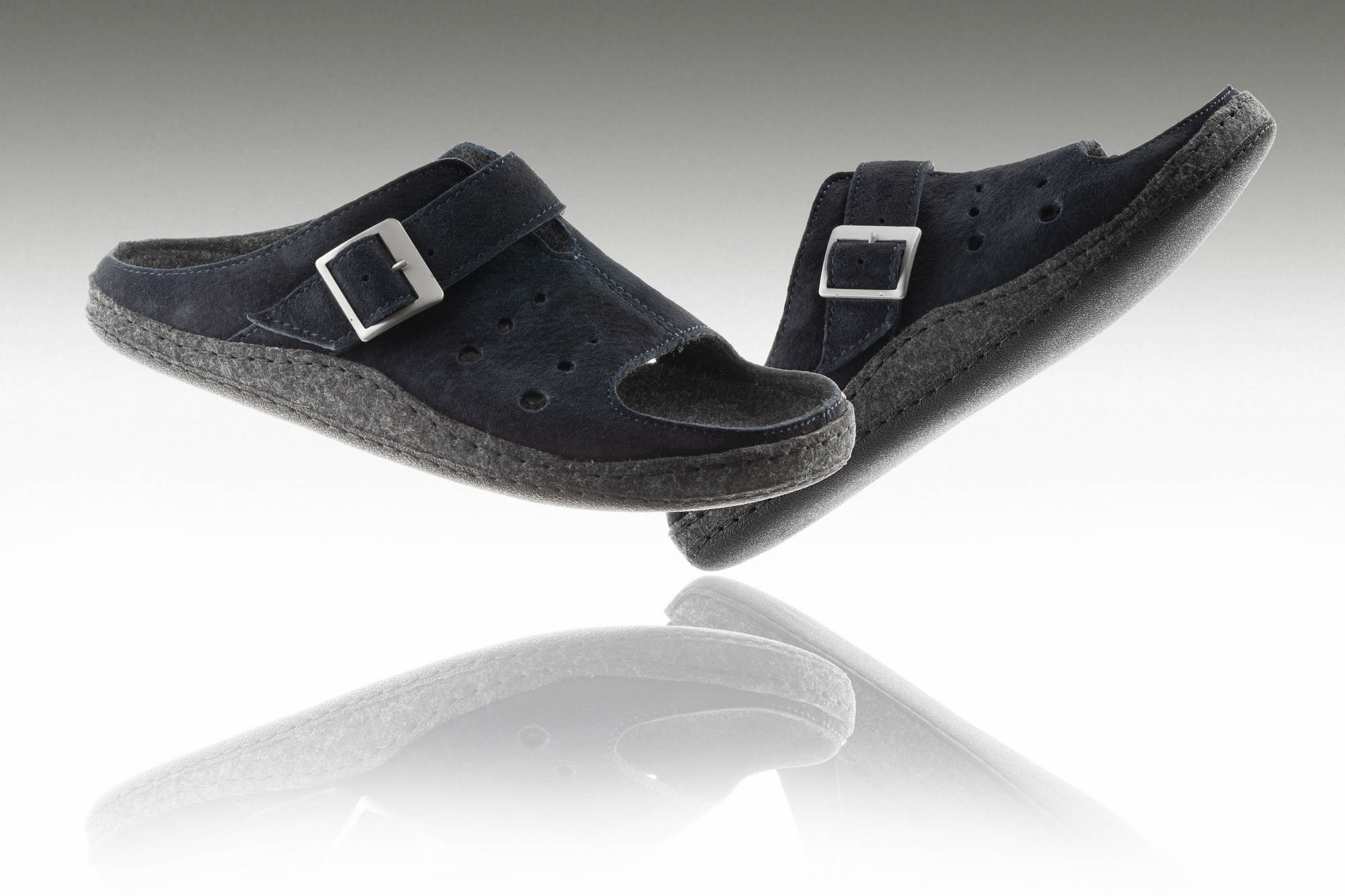 Czarne kapcie z dwoma paskami i metalowymi klamrami, uniesione nad odbiciem na białym tle.
