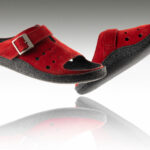 Czerwone kapcie z czarną podeszwą i regulowanym paskiem, uniesione nad odbiciem na białym tle.