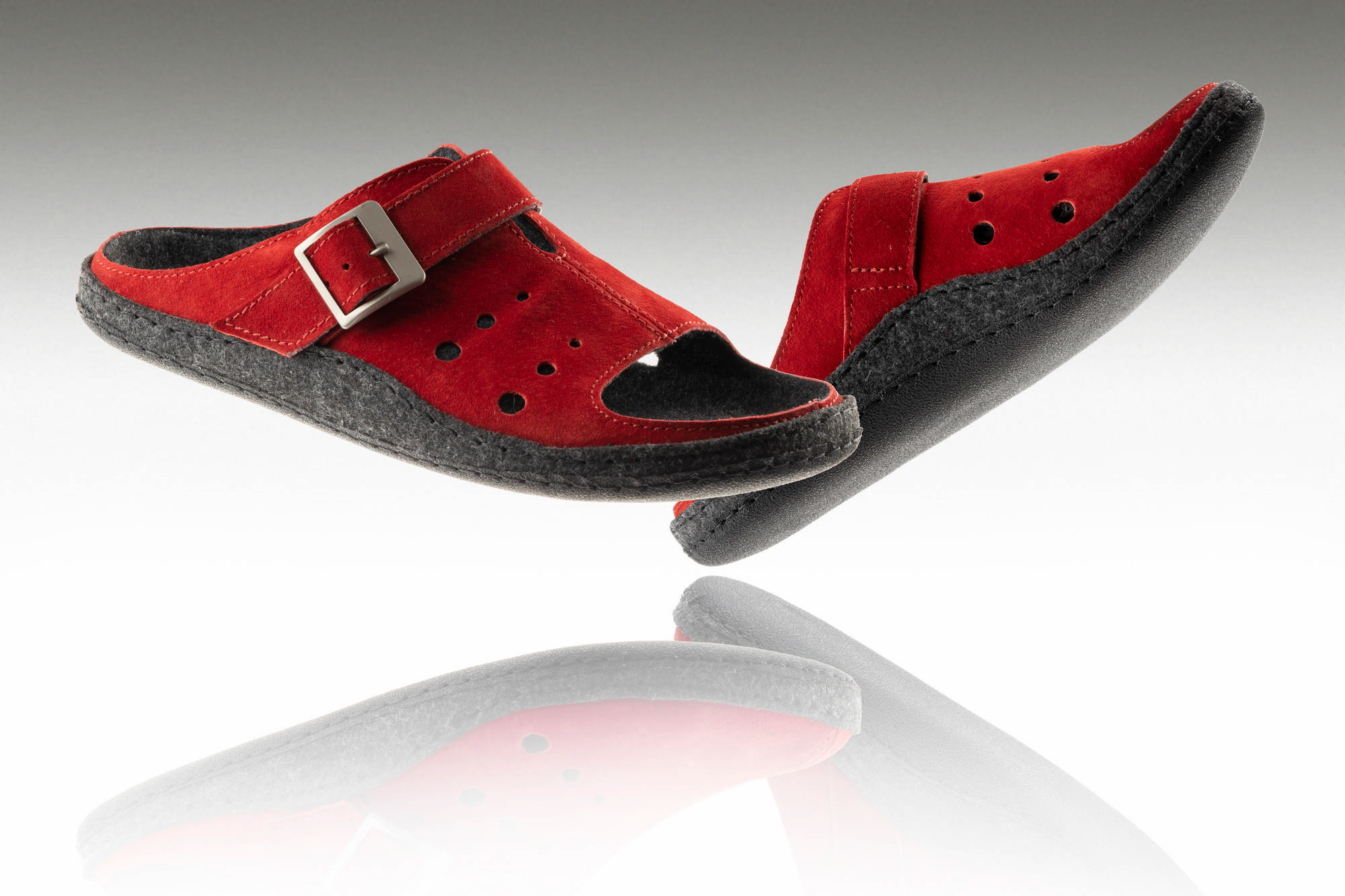 Czerwone kapcie z czarną podeszwą i regulowanym paskiem, uniesione nad odbiciem na białym tle.