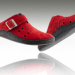 Czerwone kapcie z czarną podeszwą i regulowanym paskiem, uniesione nad odbiciem na białym tle.