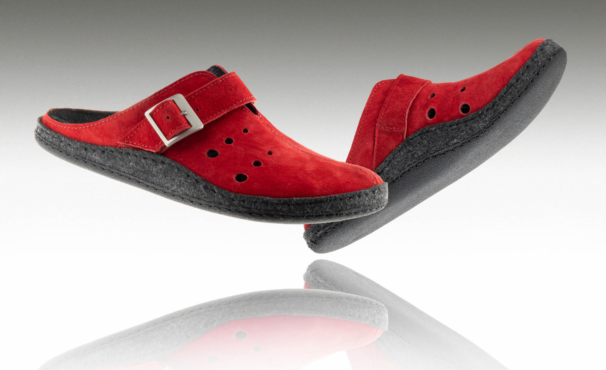Czerwone kapcie z czarną podeszwą i regulowanym paskiem, uniesione nad odbiciem na białym tle.