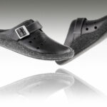 Czarne pantofle z szerokim paskiem i metalową klamrą, na podeszwie z filcu, uniesione nad odbiciem na białym tle.