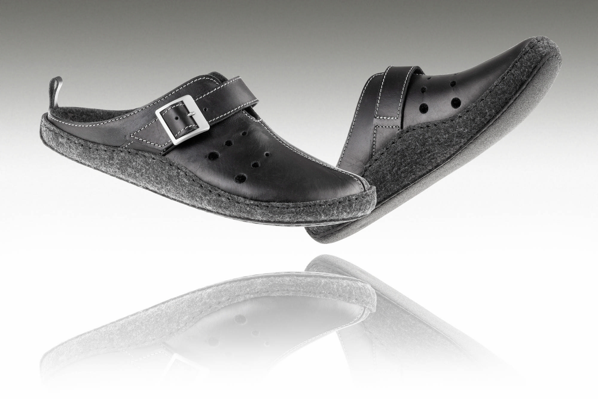 Czarne pantofle z szerokim paskiem i metalową klamrą, na podeszwie z filcu, uniesione nad odbiciem na białym tle.