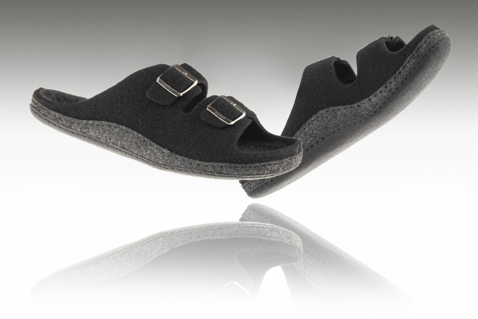 Czarne kapcie na rzepy z miękką podeszwą, uniesione nad odbiciem na białym tle.