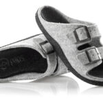 Szare kapcie z dwoma czarnymi sprzączkami, wykonane z filcu, na czarnym podbiciu, ułożone na białym tle.