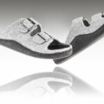 Dwie szare kapcie z dwoma paskami na rzepy, uniesione nad lustrem, na białym tle.