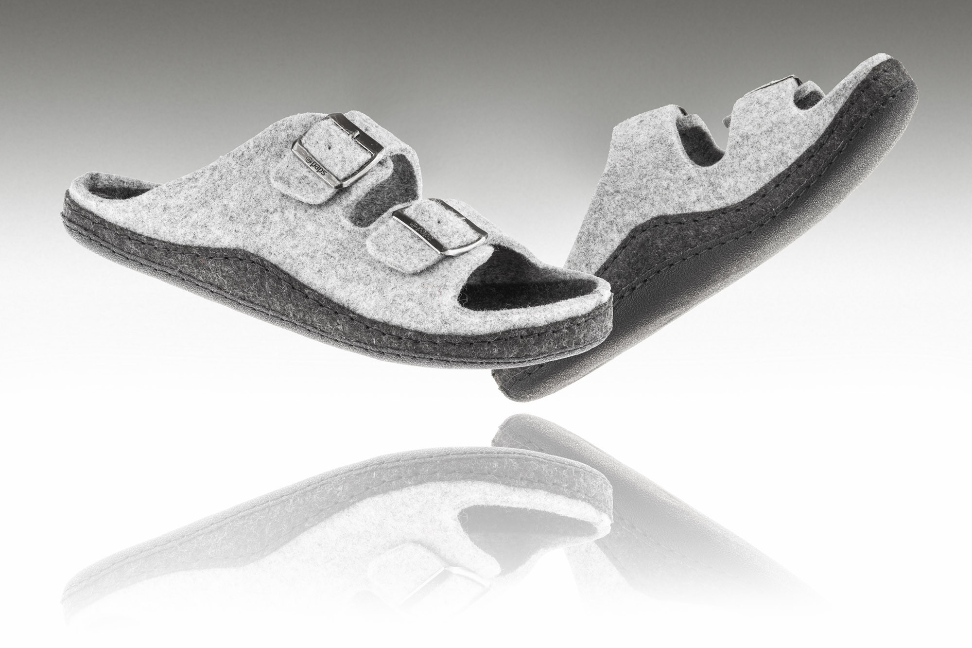 Dwie szare kapcie z dwoma paskami na rzepy, uniesione nad lustrem, na białym tle.