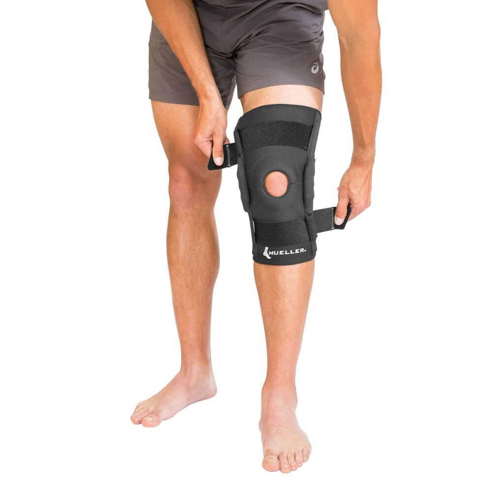 Mężczyzna zakłada na nogę stabilizator kolana z czarnego materiału, z widocznym otworem i logo MUELLER.