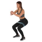 Kobieta w sportowym stroju ćwiczy przysiad z opaską na udzie i taśmą oporową, ubrana w czarne legginsy i top, na białym