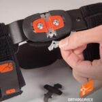 Urządzenie ortopedyczne z metalowymi elementami, osoba trzyma kluczyk do regulacji, na tle szarym.