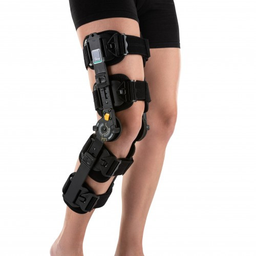 Osoba z unieruchomioną nogą w stabilizatorze ortopedycznym na udzie i podudziu, ubrana w czarne spodenki.