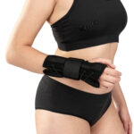 Osoba w sportowym stroju z opaską na nadgarstku, trzymająca się za brzuch, na jasnym tle.