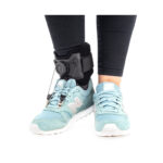 Osoba w czarnych spodniach i jasnoniebieskich butach, nosząca urządzenie wspomagające chodzenie z czujnikiem na kostce.