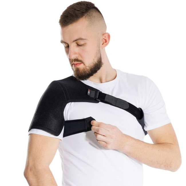 Mężczyzna zakładający ortozę na ramię, ubrany w biały t-shirt, z zamkniętymi oczami, na białym tle.