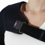 Osoba nosi czarny ochraniacz na ramię z logo SPORTS i regulowanym paskiem, na białej koszulce.