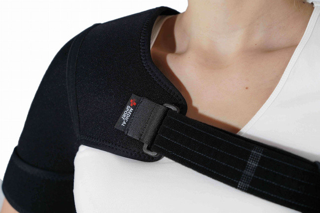 Osoba nosi czarny ochraniacz na ramię z logo SPORTS i regulowanym paskiem, na białej koszulce.