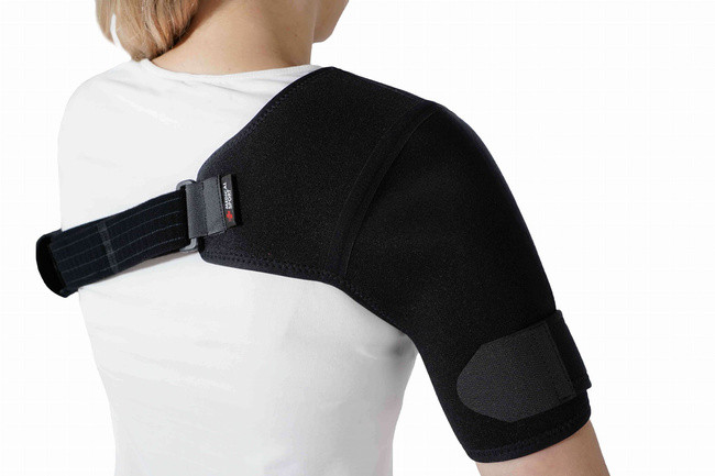 Osoba nosząca czarny stabilizator na ramieniu, z regulowanym paskiem na piersi, w białej koszulce.