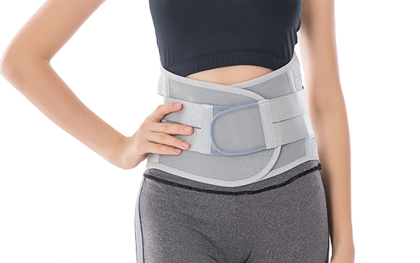 Osoba nosząca biały pas ortopedyczny na talii, ubrana w czarny top i szare legginsy.