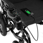 Elektryczny wózek inwalidzki z ładowarką i telefonem na podłodze, pokazującym ładowanie baterii.