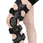 Orteza na kolanie z czarnymi paskami i elementami, mocowana na nogę, służy do stabilizacji i wsparcia stawu kolanowego.