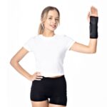 Uśmiechnięta kobieta w białym t-shircie i czarnych szortach, pokazuje dłoń w rękawicy sportowej na tle białym.