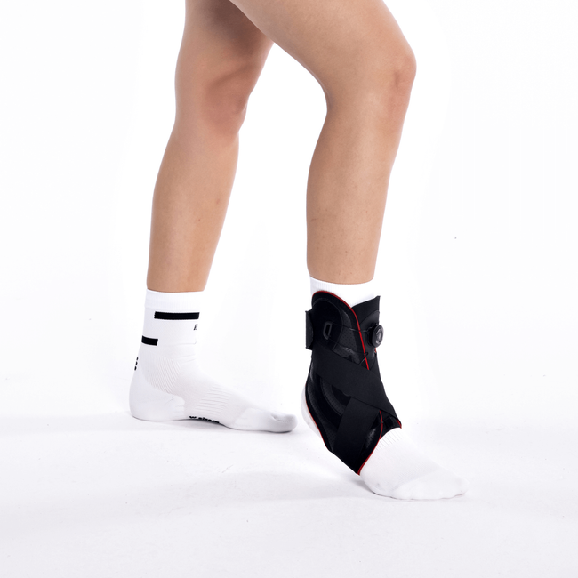 Stopa z ortezą na kostce, druga stopa w białych skarpetach, osoba stoi na białym tle.