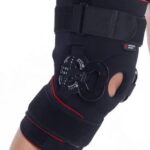 Orteza kolana z mechanizmem rotacyjnym i paskiem, czarna z czerwonymi akcentami, zapewniająca stabilizację i wsparcie.