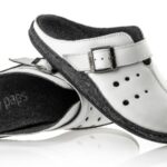 Białe kapcie z czarną podeszwą, zapinane na regulowany pasek z klamrą, przeznaczone do użytku domowego.