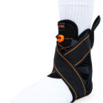 Orteza stopy z czarnym opaskiem, zapięciem na rzep i pomarańczowym elementem, zakładana na stopę w białym skarpecie.