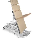 Stół do badania ginekologicznego z regulowanym oparciem i podnóżkiem, na metalowej podstawie z kółkami.