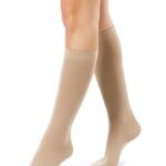 Kobieta w beżowych pończochach sięgających do kolan, stojąca na białym tle.