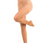 Kobieta w beżowych pończochach, stoi na jednej nodze, z drugą lekko uniesioną, na białym tle.