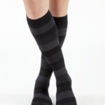 Nogi kobiety w czarno-szarych, długich, wzorzystych skarpetach z poziomymi pasami, stojącej na białym tle.