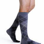 Mężczyzna nosi długie skarpetki w szary i czarny wzór argyle, sięgające do kolan, na białym tle.