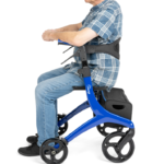Osoba starsza na specjalnym wózku inwalidzkim z podnóżkiem i torbą pod siedzeniem, ubrana w koszulę i jeansy.